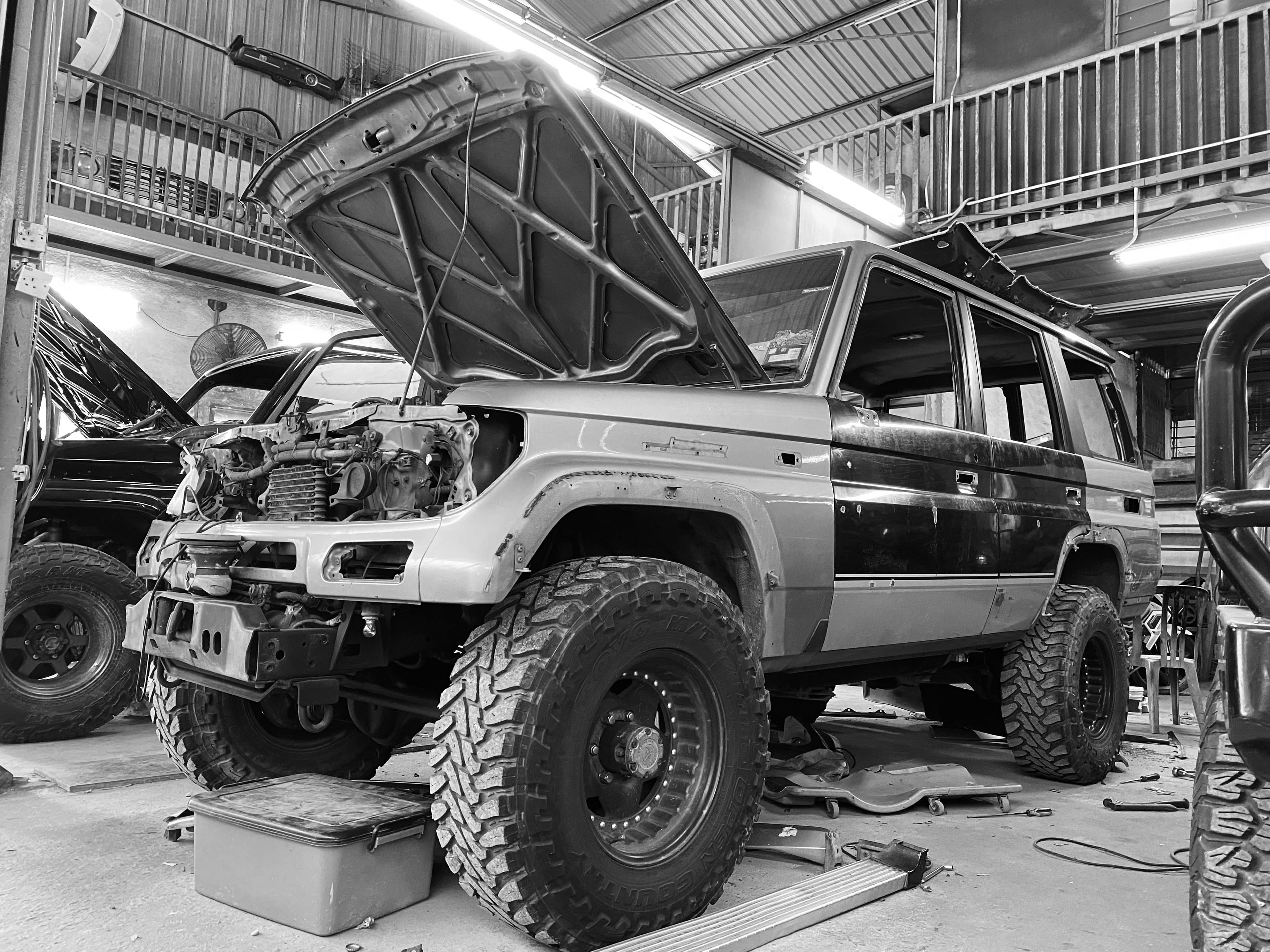 A Land Cruiser mid-rebuild in a workshop, bonnet up and engine bay open, on mud-terrain tyres