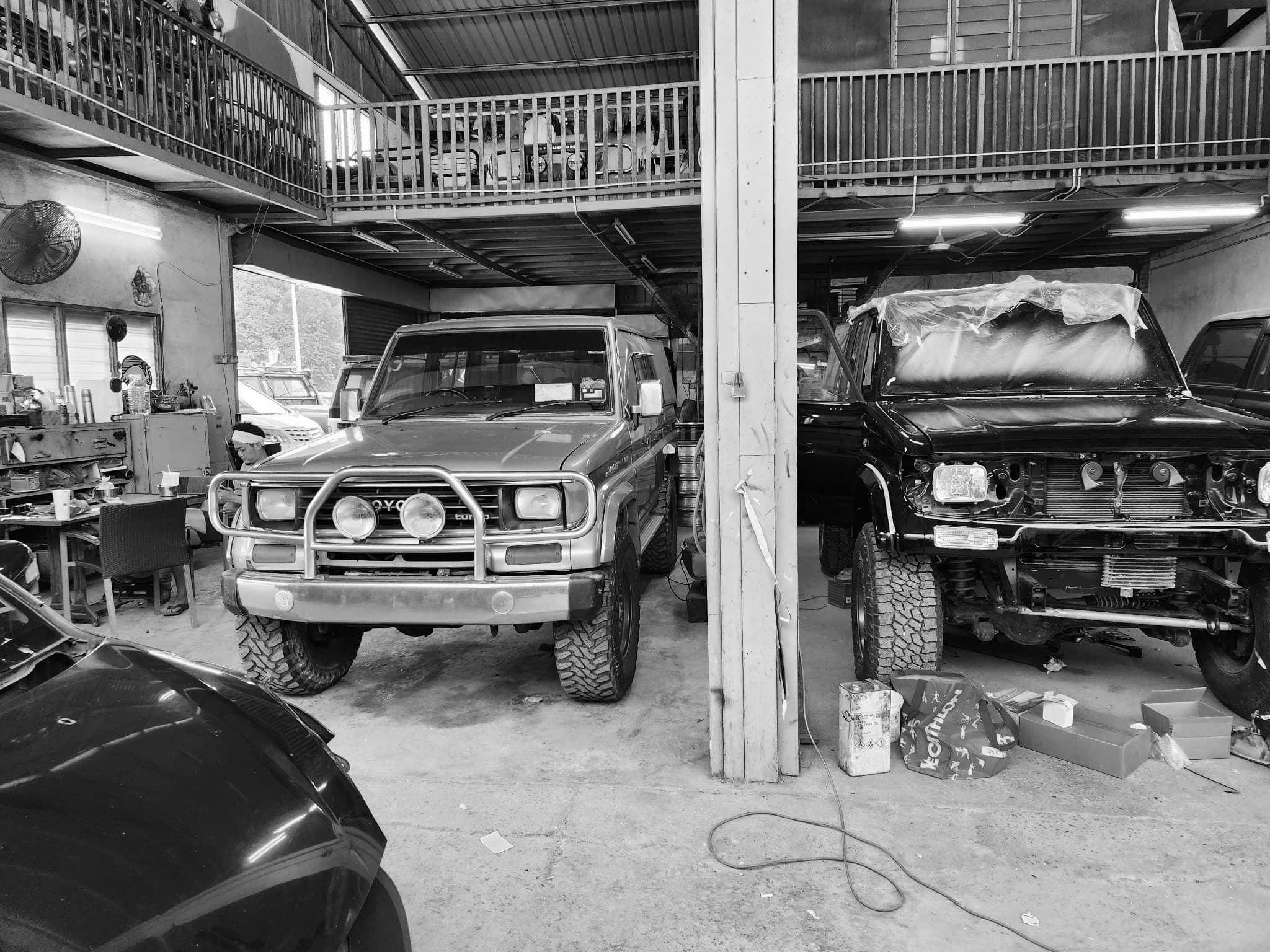 Two Toyota Land Cruisers parked in a workshop under a mezzanine, one mid-repair with its bonnet off